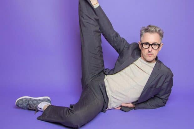 Alan Cumming wearing glasses and stretching one leg while lying on a purple floor, HD Desktop Wallpaper
