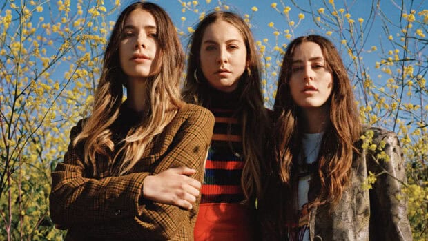 Alana Haim Wallpapers show three women standing among yellow flowers under a clear blue sky, HD Desktop Wallpaper