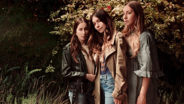 Alana Haim and her sisters are standing together near foliage wearing casual outfits, HD Desktop Wallpaper