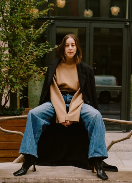 Alana Haim wears a tan sweater with jeans and black boots sitting on a bench, HD Mobile Wallpaper