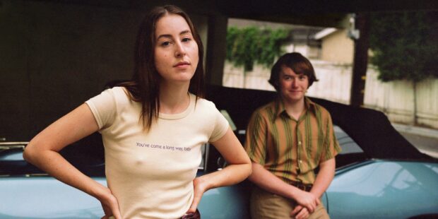 Alana Haim stands confidently near a blue car with a person sitting in the background, 2K Desktop Wallpaper