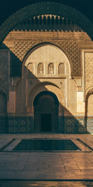 The image shows ancient Moroccan architecture with detailed carvings and mosaic tiles around a small fountain, HD Mobile Wallpaper