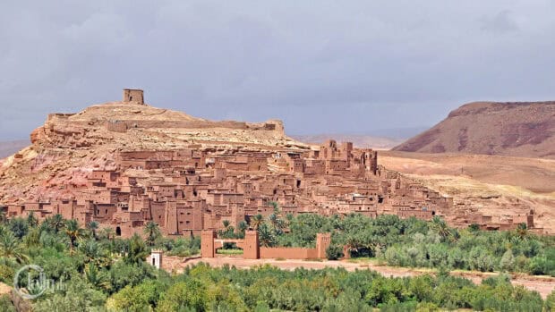 An ancient village with traditional clay buildings stands amid greenery under a cloudy sky in Morocco, 4K Desktop Wallpaper