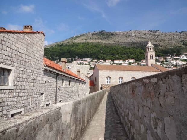 A stone pathway runs along the ancient city walls in Dubrovnik with buildings and hills in the background, 2K Desktop Wallpaper