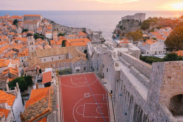 The ancient city walls in Dubrovnik surround a red basketball court near the sea at sunset, HD Desktop Wallpaper