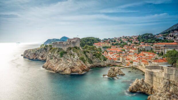The ancient city walls of Dubrovnik stretch along the rugged coastline beneath a cloudy sky, 4K Desktop Wallpaper