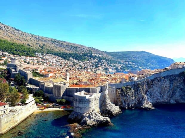 The ancient city walls of Dubrovnik stretch along the rocky coastline under a clear blue sky, HD Desktop Wallpaper
