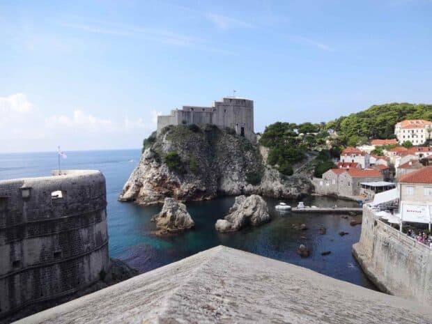 The ancient city walls of Dubrovnik rise above the calm sea with rocky shores and red roofed buildings, 2K Desktop Wallpaper
