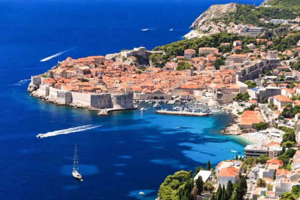 The ancient city walls of Dubrovnik overlook the bright blue sea and boats near the coast, 2K Desktop Wallpaper