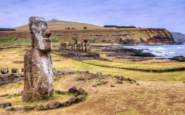 A large stone statue stands on the grassy terrain near a coastal cliff with more statues in the background, 2K Desktop Wallpaper