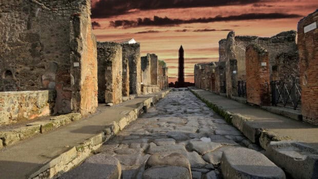 The ancient cobblestone street in Pompeii shows ruins under a dramatic sunset sky, 2K Desktop Wallpaper