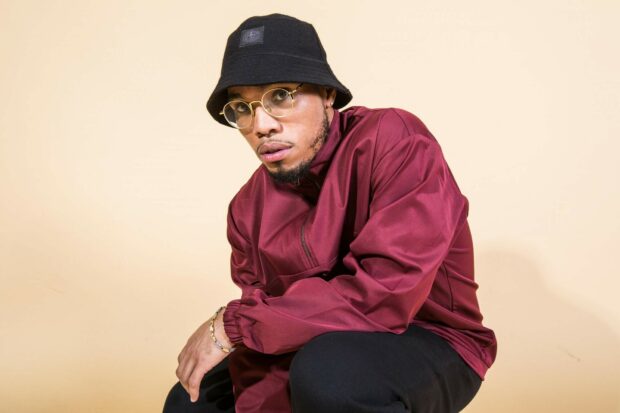 Anderson Paak is wearing a maroon jacket and black bucket hat posing against a beige background, HD Desktop Wallpaper