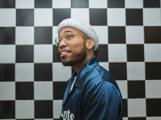 Anderson Paak wears glasses and a knitted hat smiling against a checkered wall, HD Desktop Wallpaper