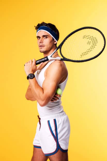 Antoni Porowski holds a tennis racket while wearing a sporty outfit against a yellow background, 2K Mobile Wallpaper