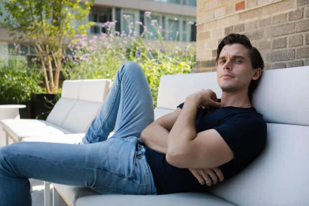 Antoni Porowski is sitting relaxed on a white outdoor couch with greenery in the background, 2K Desktop Wallpaper