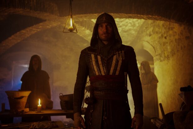A man in a hooded cloak stands in an ancient chamber with candles and shadows, 2K Desktop Wallpaper
