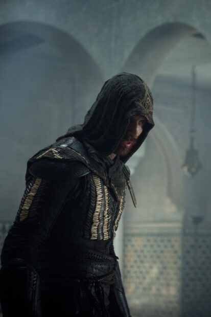 A hooded assassin from the Assassins Creed movie stands quietly in a dimly lit ancient setting, 2K Mobile Wallpaper