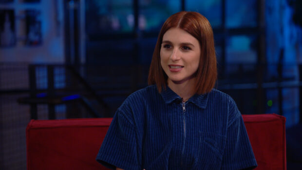 Aya Cash is sitting on a red sofa wearing a blue shirt in a dimly lit room, HD Desktop Wallpaper