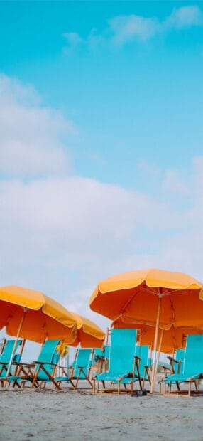 Bright orange beach umbrellas with blue chairs on sandy shore under a clear sky, 2K Phone Wallpaper