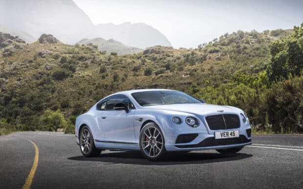 A silver Bentley Continental GT is parked on a winding mountain road with greenery and hills in the background, 2K Desktop Wallpaper