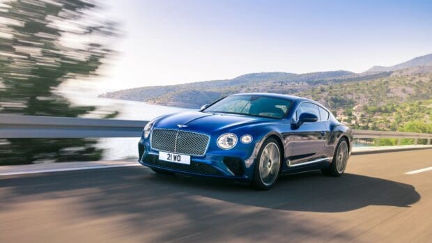 A blue Bentley Continental GT is driving along a scenic coastal highway with hills and water in the background, 4K Desktop Wallpaper