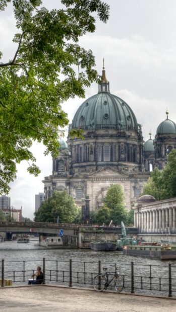 A serene view of Berlin Cathedral dome by the riverside with trees and a bicycle near the water, HD Mobile Wallpaper