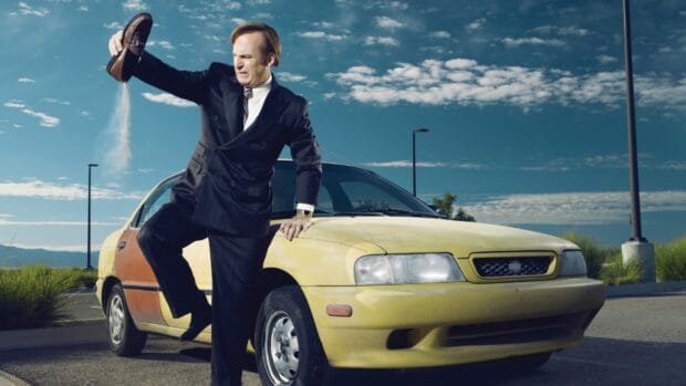 A man in a suit throwing sand from his shoe near a yellow car under a blue sky with clouds, HD Desktop Wallpaper