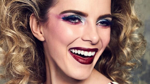 Betty Gilpin Wallpapers shows a smiling woman with vibrant eye shadow and glitter, 4K Mobile Wallpaper