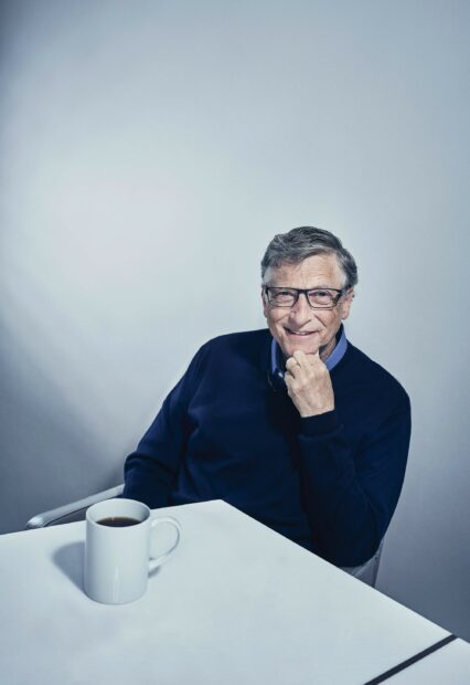 Bill Gates thoughtful pose wearing glasses and a dark sweater sitting at a white table with a coffee mug, 2K Mobile Wallpaper