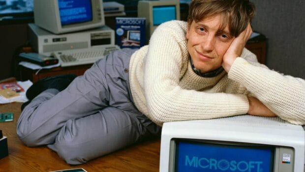 Bill Gates is lying next to old Microsoft computer screens in a retro office setting, HD Desktop Wallpaper