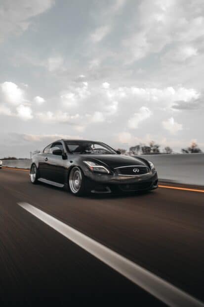 A black Infiniti G37 is driving fast on a road with blurred surroundings and cloudy sky, 2K Mobile Wallpaper