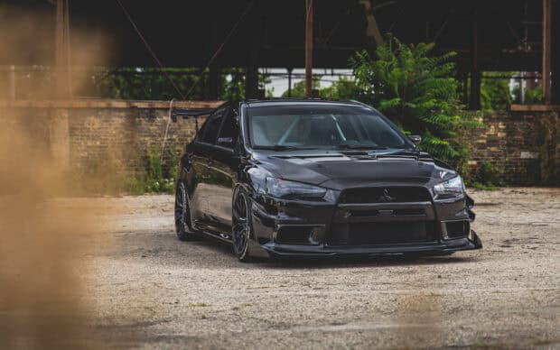 A black Lancer Evo is parked outdoors near a brick wall and greenery, showcasing its sleek design, 2K Desktop Wallpaper