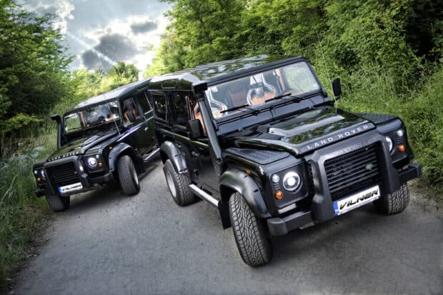 A black Land Rover Defender vehicle parked on a narrow road surrounded by green trees and bushes, 2K Desktop Wallpaper