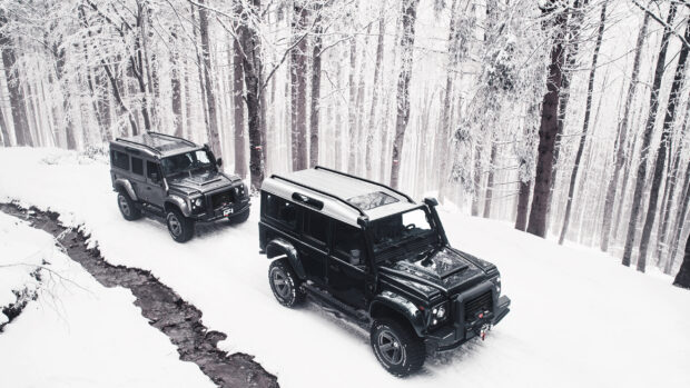 Two Land Rover Defender vehicles drive through a snowy forest with tall trees and a small stream, 2K Desktop Wallpaper