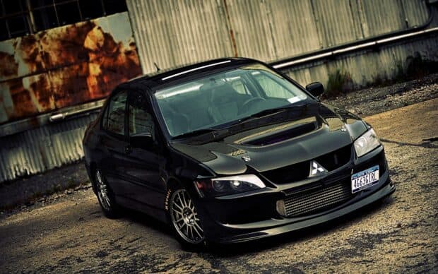 A black Mitsubishi Lancer Evo is parked on a rough concrete surface near a rusty corrugated metal wall, HD Desktop Wallpaper