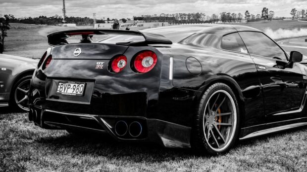 A black Nissan GT R car is parked outdoors with a blurred motorsport park in the background, HD Desktop Wallpaper