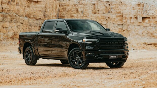 A black Ram pickup truck is parked on a sandy desert terrain with rocky cliffs in the background, 2K Desktop Wallpaper