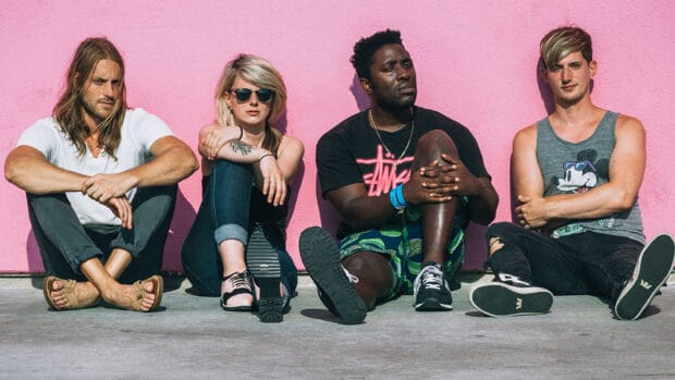 The Bloc Party band members are sitting outdoors against a pink wall in casual outfits, 2K Desktop Wallpaper