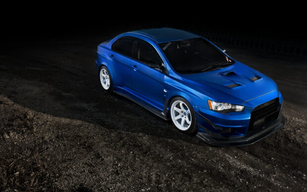 A blue Lancer Evo with white wheels parked on a dark gravel surface, 2K Desktop Wallpaper