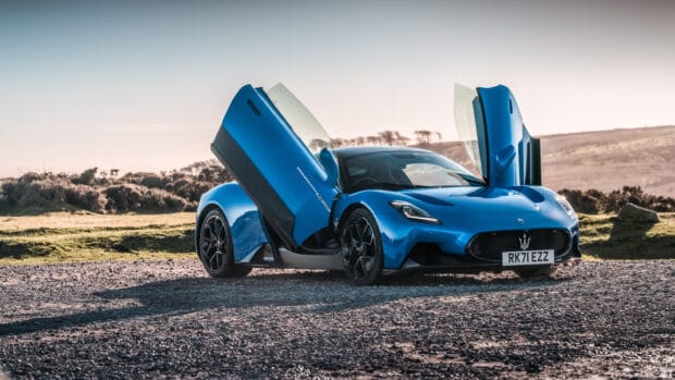 A blue Maserati MC20 is parked with its scissor doors open in a natural outdoor setting, 2K Desktop Wallpaper