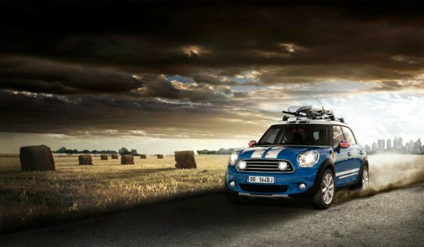 A blue Mini Cooper drives on a rural road with hay bales and stormy clouds, HD Desktop Wallpaper