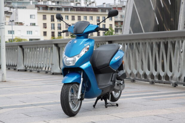 A blue Peugeot Kisbee scooter is parked on a city bridge with buildings in the background, HD Desktop Wallpaper