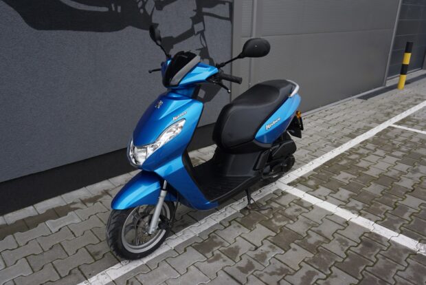A blue Peugeot Kisbee scooter is parked on a paved parking area near a gray wall, HD Desktop Wallpaper