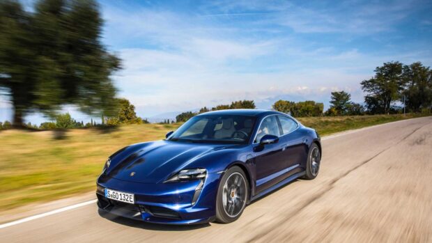 A blue Porsche Taycan is driving quickly on a scenic road surrounded by trees and grass under a clear sky, HD Desktop Wallpaper