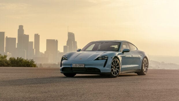 A blue Porsche Taycan is parked on empty ground with city buildings visible in the background during sunset, 4K Desktop Wallpaper