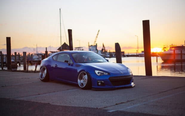 A blue Subaru Brz is parked by the harbor with boats in the background during sunset, 2K Desktop Wallpaper