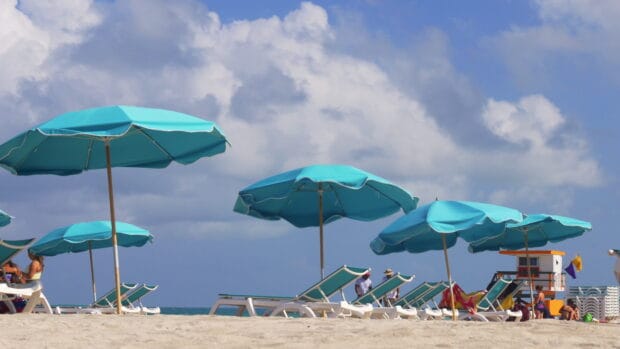 Several blue beach umbrellas are set up on a sandy shore with lounge chairs and people enjoying the beach, 4K Desktop Wallpaper