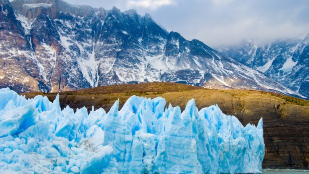 The glacier shows bright blue ice details beneath rugged brown mountains, 4K Desktop Wallpaper