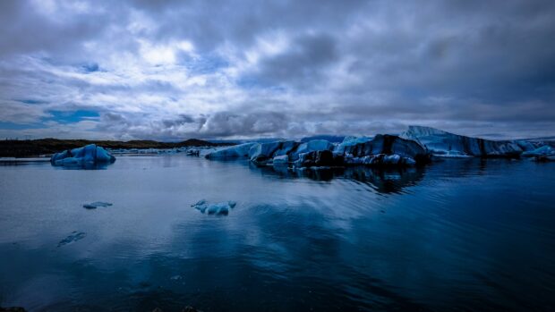 The glacier ice floats peacefully on the clear water reflecting the cloudy sky above in a cold environment, 4K Desktop Wallpaper