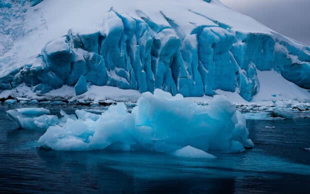 A blue glacier with floating ice chunks sits in cold water under a gray sky, 2K Desktop Wallpaper